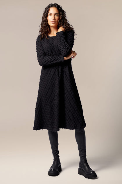 A woman wearing a luxurious texture black Pocket Jersey Flared Dress and soft jersey leggings.