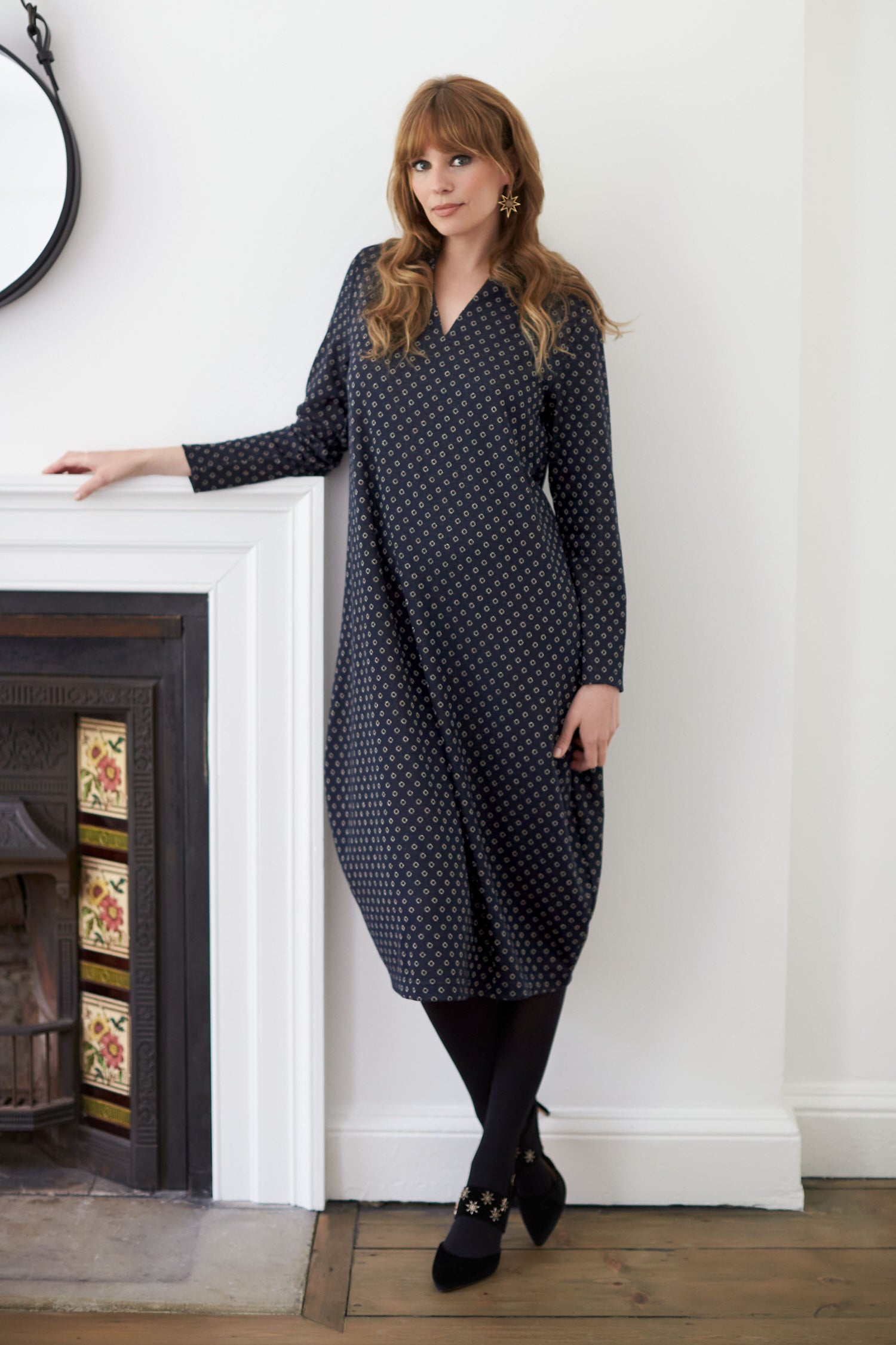 A woman in a Lurex Spot Jersey Dress leaning against a fireplace.
