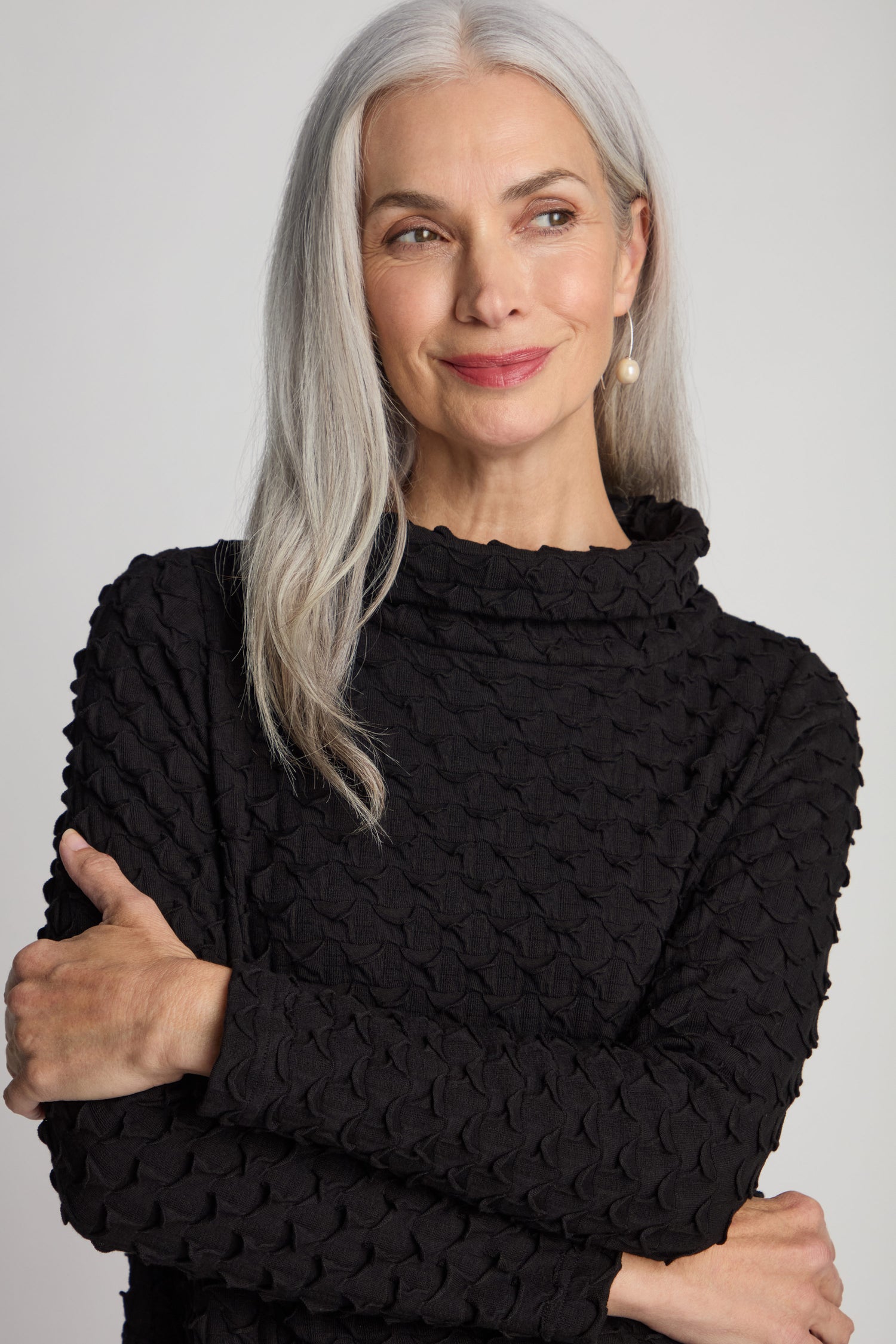 A woman with long gray hair smiles with her arms crossed, wearing the Pocket Jersey Cowl Neck Dress in a textured black fabric.