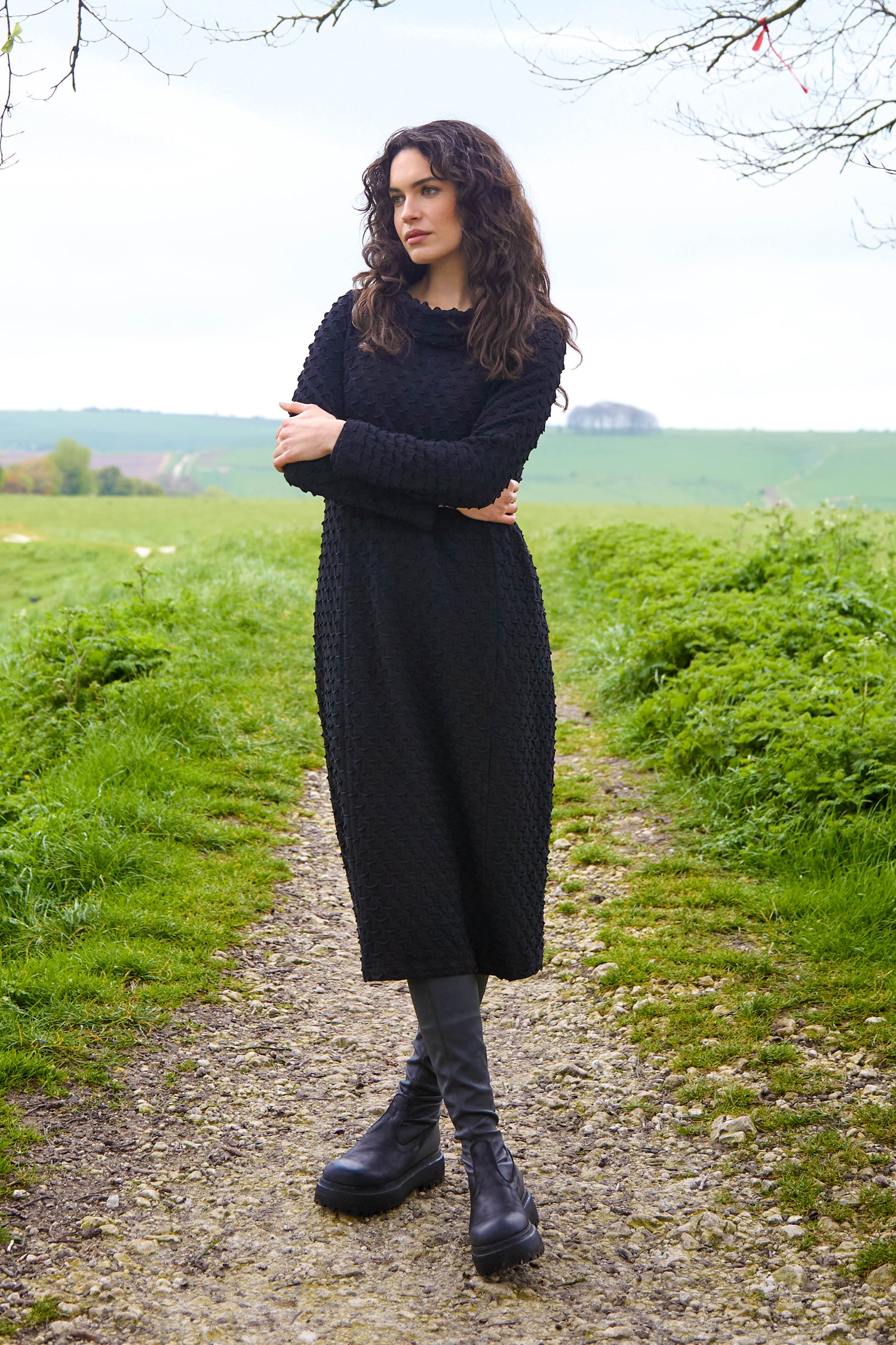 A person in a slim silhouette, wearing the Pocket Jersey Cowl Neck Dress in black with boots, stands on a dirt path in a grassy landscape.