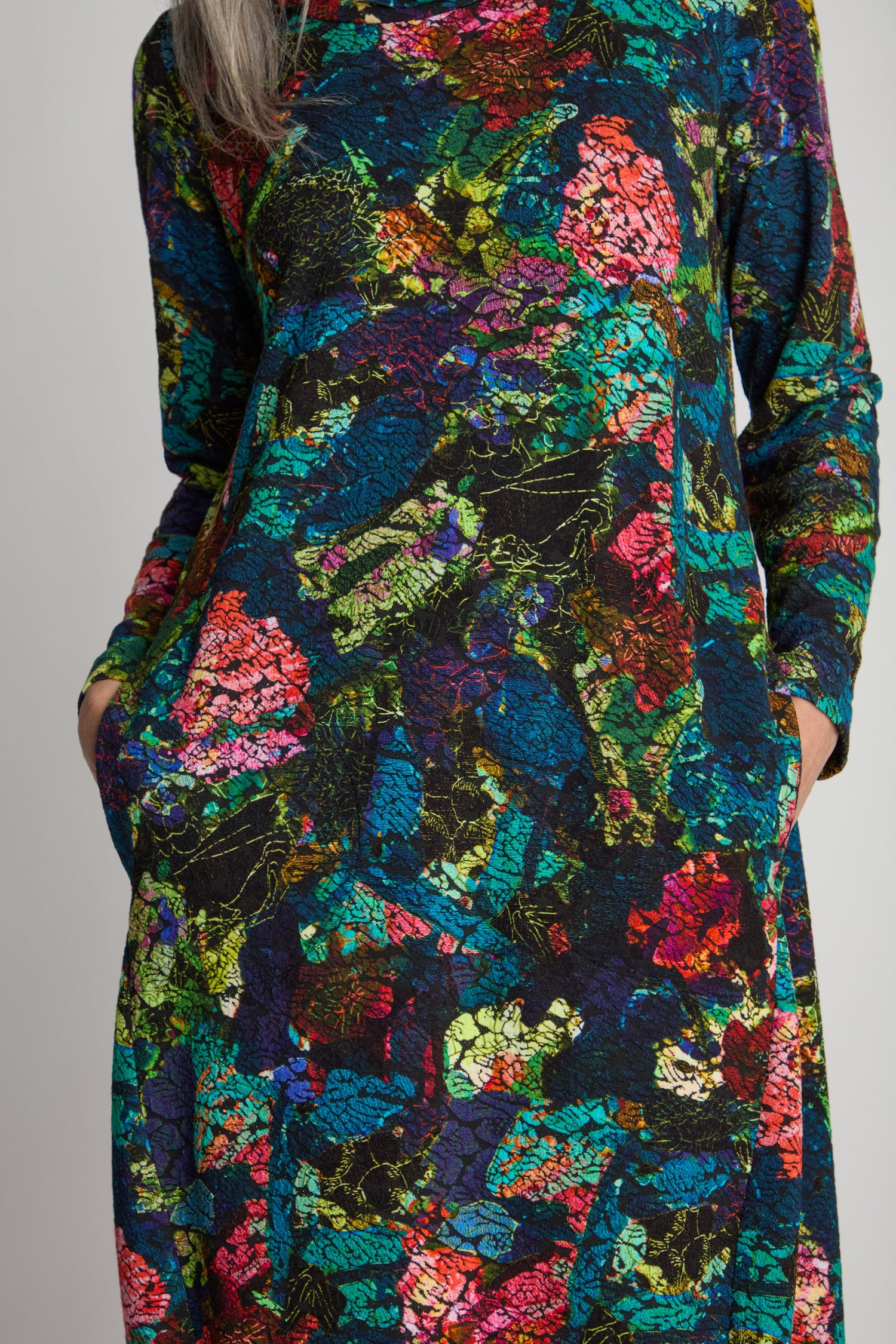 A person wearing a vibrant Abstract Floral Jersey Bubble Dress, adorned with a colorful abstract pattern of pink, blue, green, and yellow splashes, featuring long sleeves and a convenient pocket.