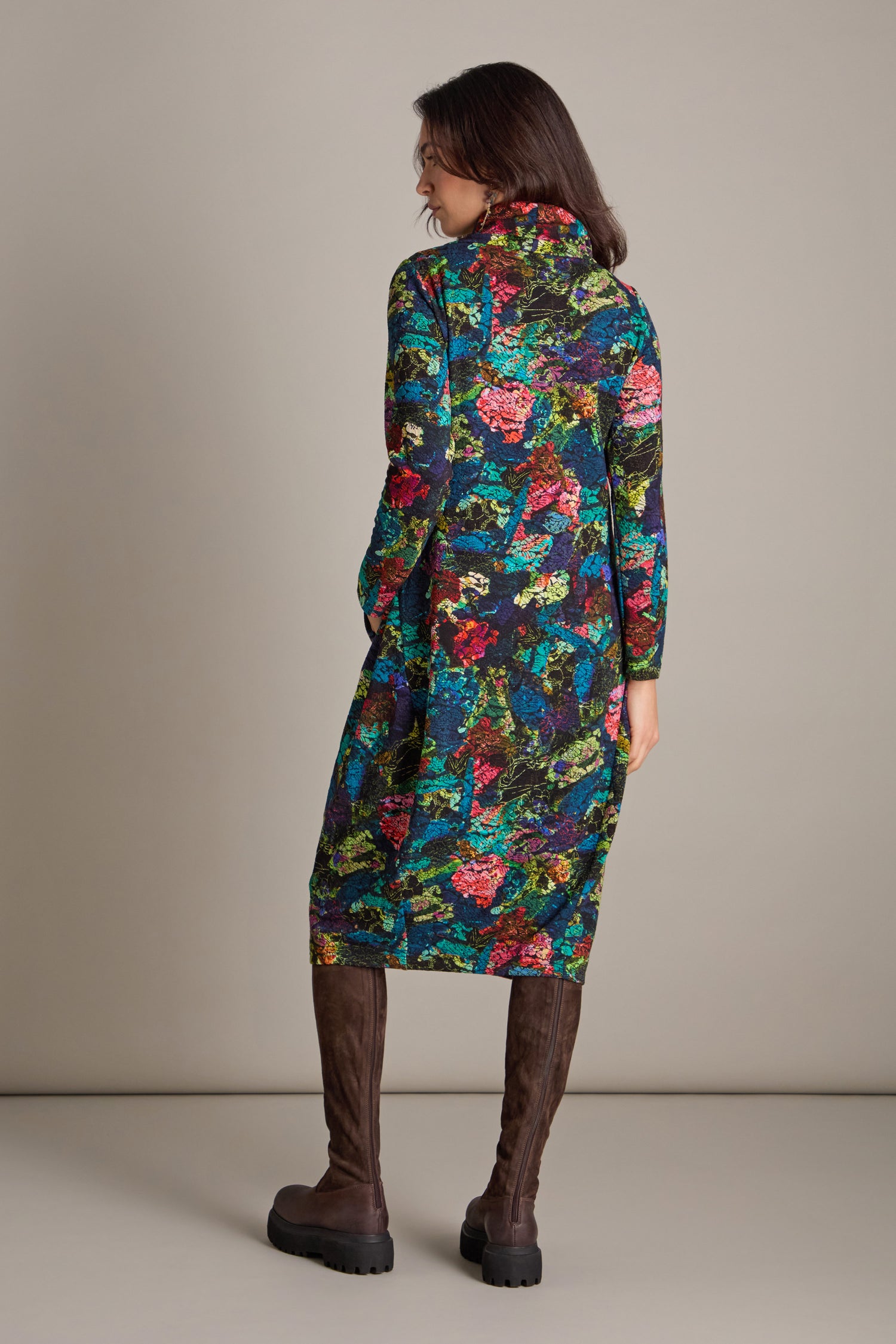 A person wearing a long, Abstract Floral Jersey Bubble Dress and tall brown boots stands facing away against a plain background.