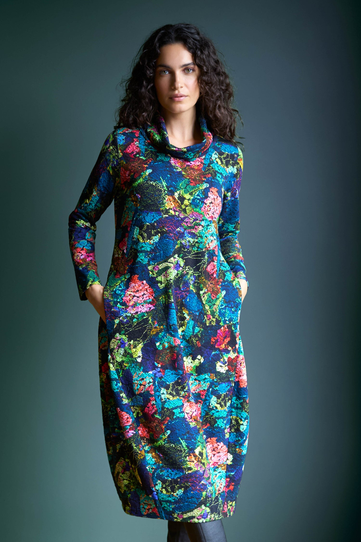 Dressed in the long-sleeve Abstract Floral Jersey Bubble Dress with a cowl neckline, a person stands gracefully against a dark green background.