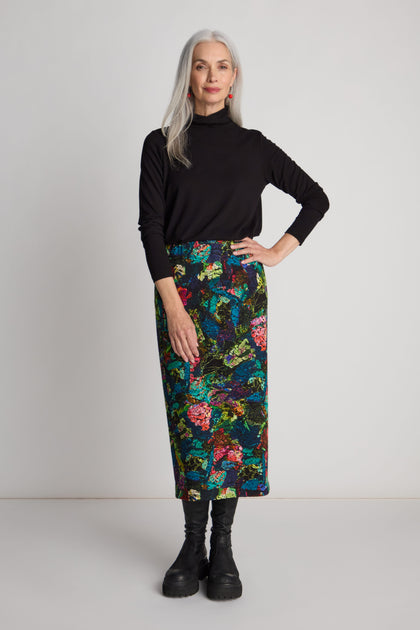 A woman with long white hair, wearing a black turtleneck, the Abstract Floral Jersey Tube Skirt with an elasticated waistband, and black boots, stands against a plain background.