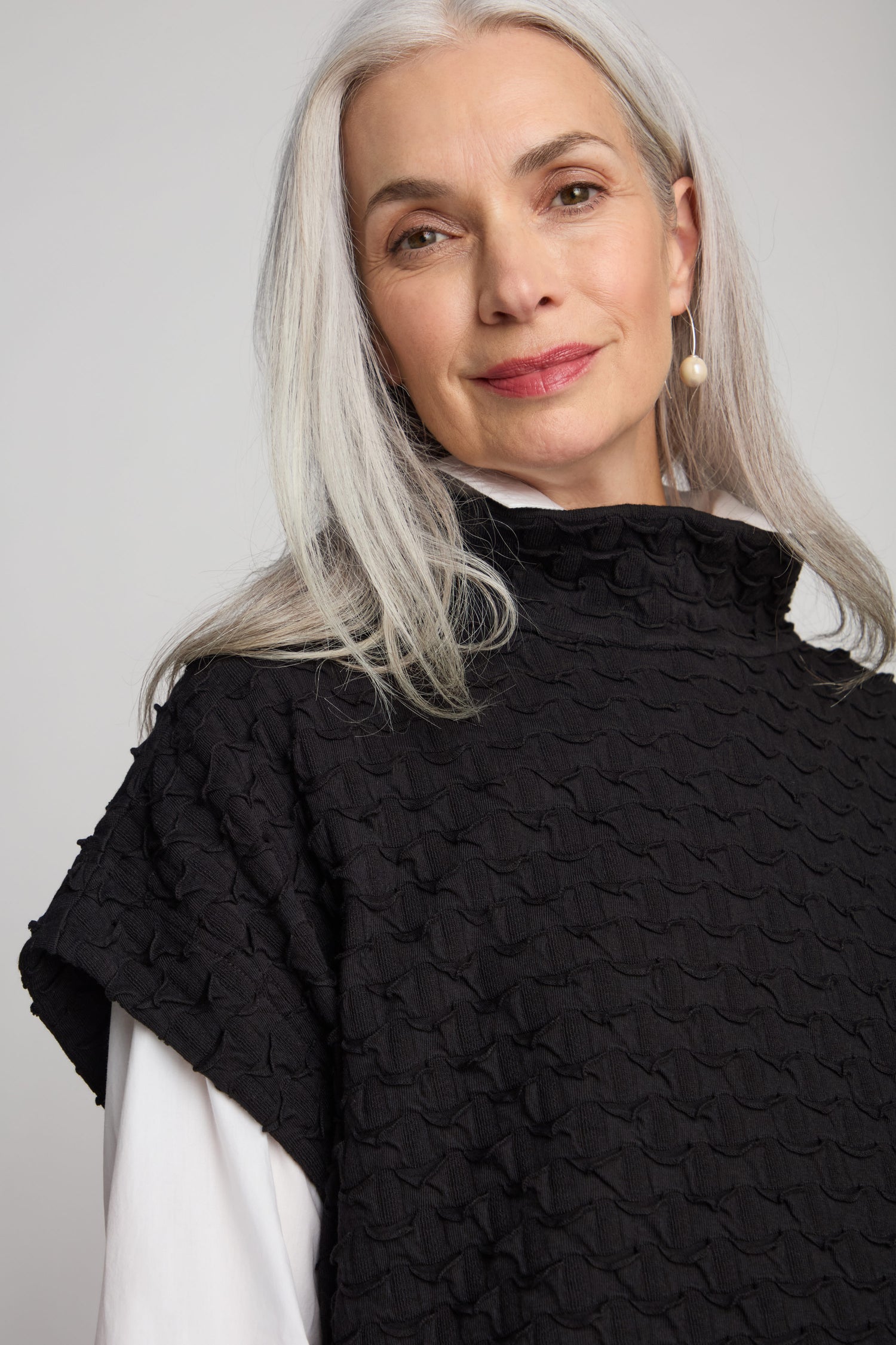 A woman with long gray hair, dressed in a luxurious black Pocket Jersey Tabard and pearl earrings, smiles slightly at the camera against a plain background.