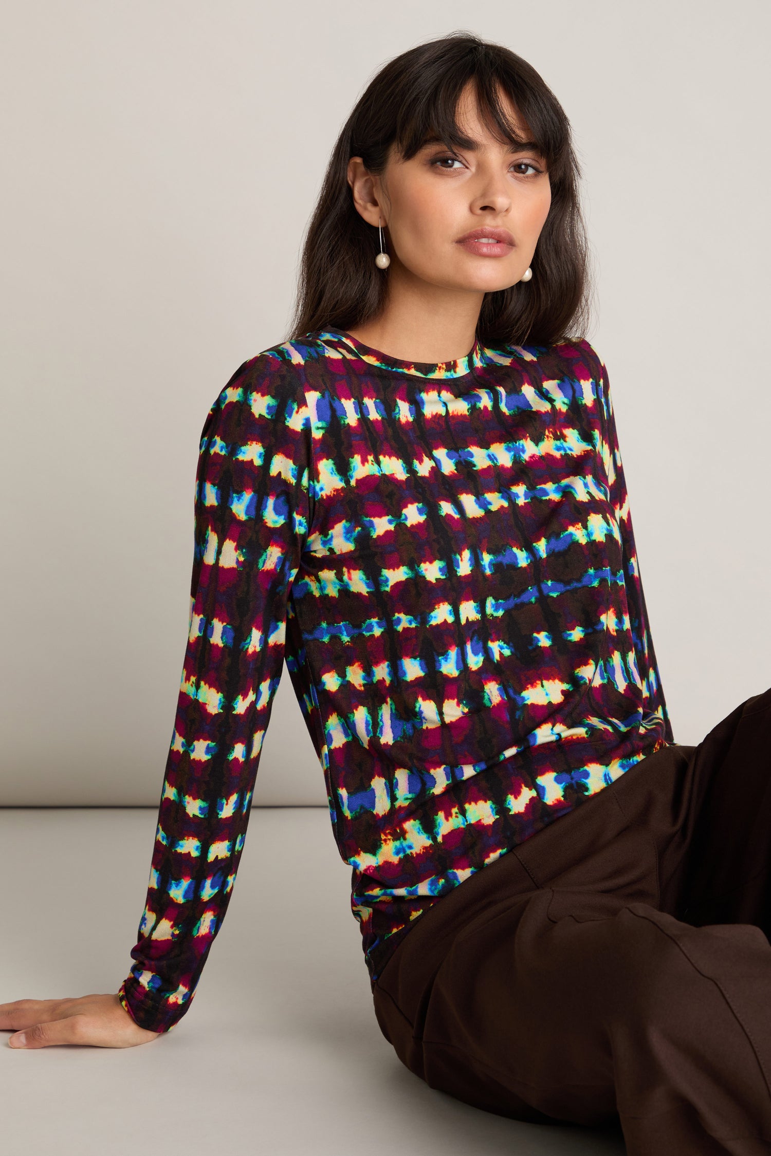 A person with long dark hair wearing the Marble Stripe Jersey Top and dark pants sits on the floor against a plain background, highlighting why this colorful patterned top has become such a popular layering piece.