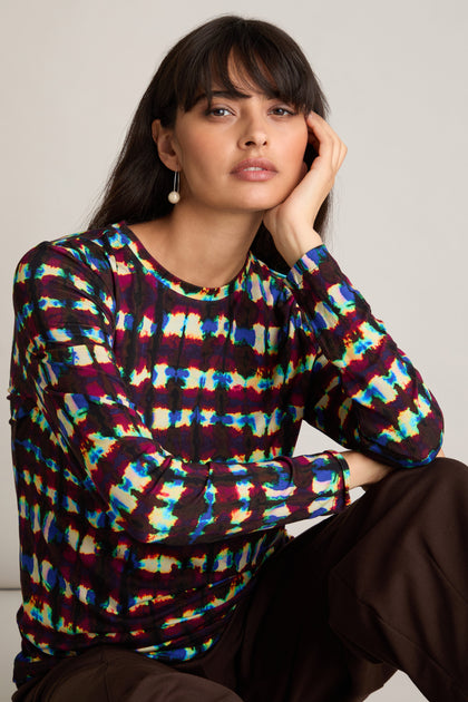 A woman in a Marble Stripe Jersey Top featuring a classic crew neckline paired with brown pants, resting her face on her hand and gazing towards the camera.