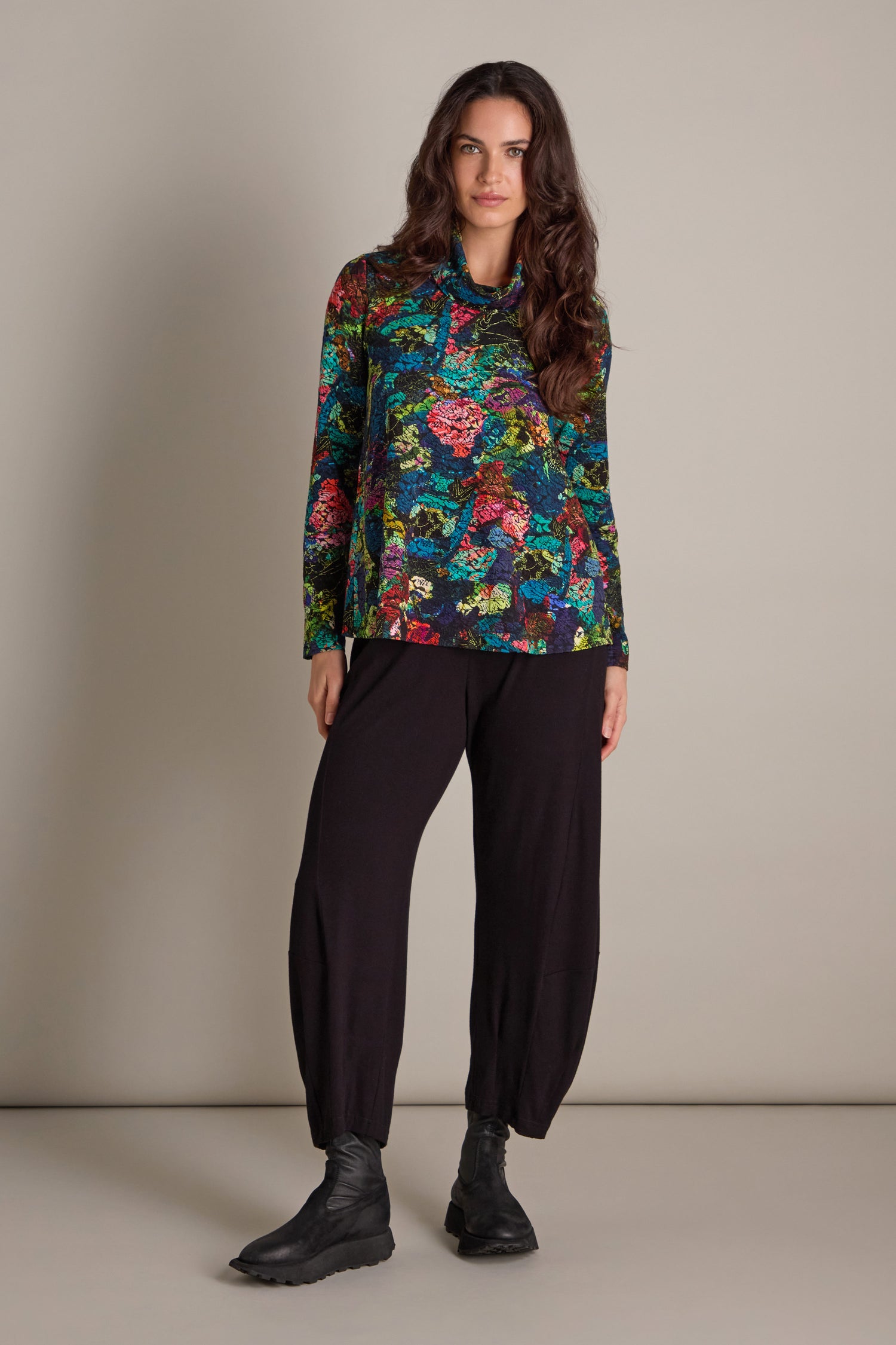 A person stands in front of a plain background wearing the Abstract Floral Jersey Cowl Neck Top, paired with black pants and black shoes.