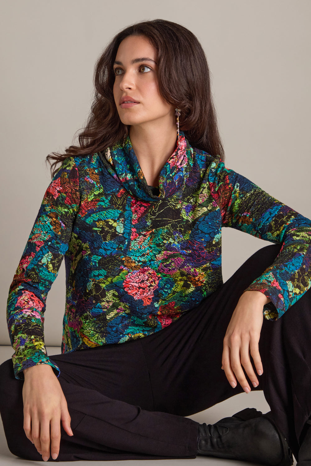 A person sits cross-legged, draped in an Abstract Floral Jersey Cowl Neck Top paired with black pants, gazing thoughtfully to the side.