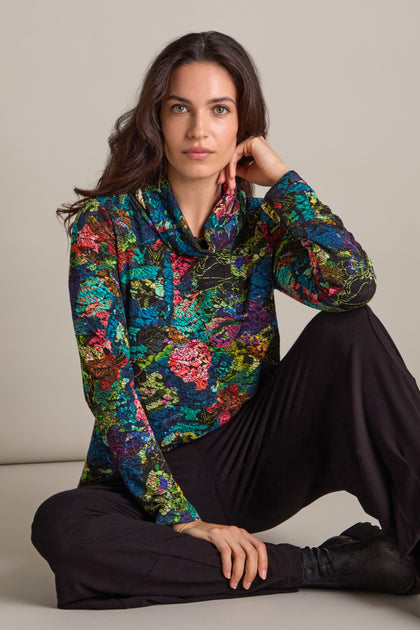 A person with long dark hair sits on the floor, dressed in an Abstract Floral Jersey Cowl Neck Top and black pants. Their vibrant attire is akin to the Sahara Exclusive collection and stands out against a plain background.