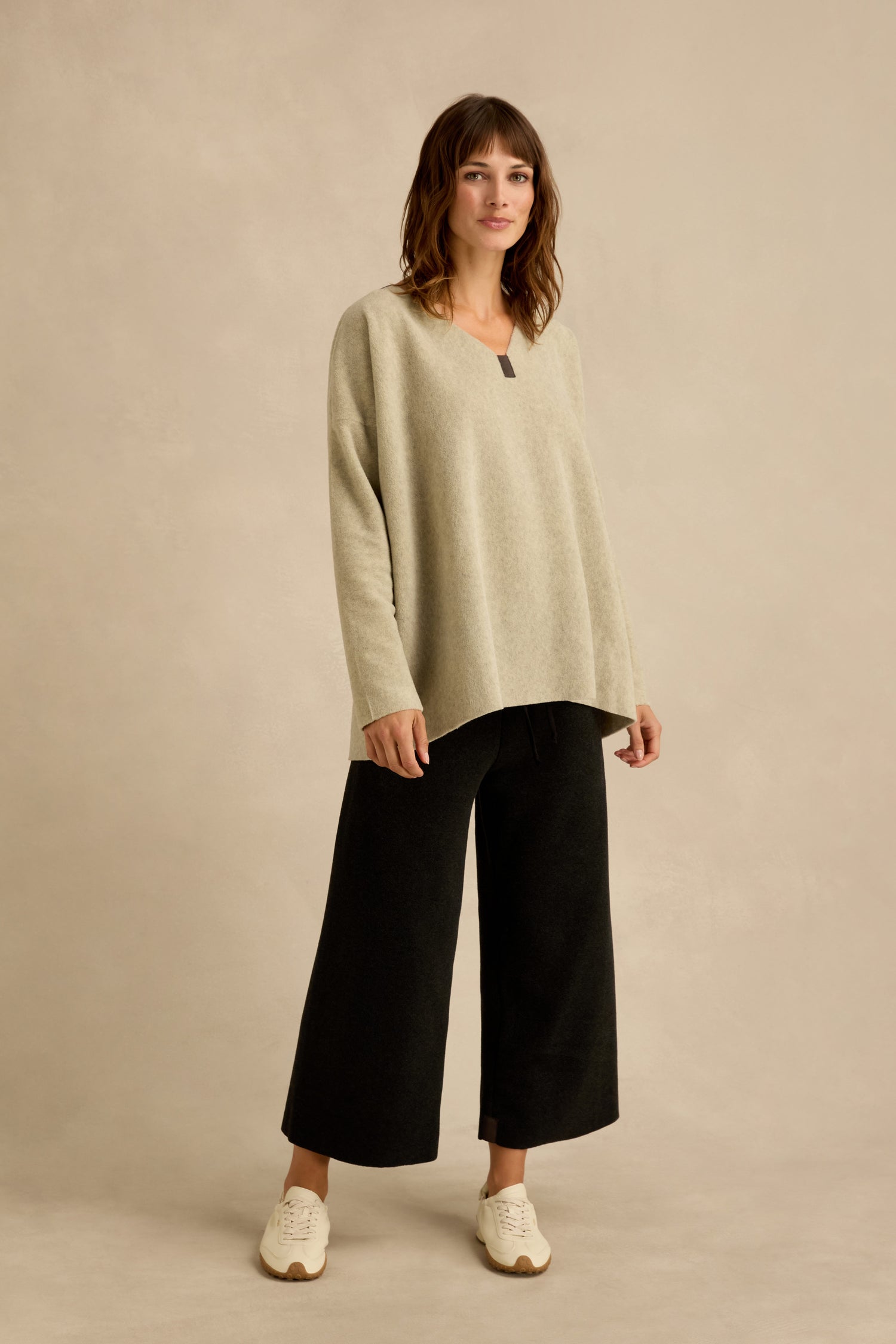 A woman wearing a loose Cream Marl V Neck Fleece Tunic, wide-leg black pants, and white sneakers stands against a neutral background.