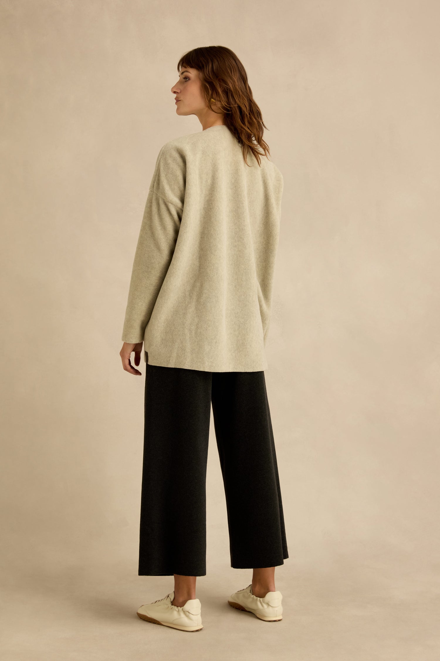 A person stands facing away, wearing a Cream Marl V Neck Fleece Tunic from Henriette Steffensen, paired with wide-leg black pants and white sneakers, against a plain neutral background.