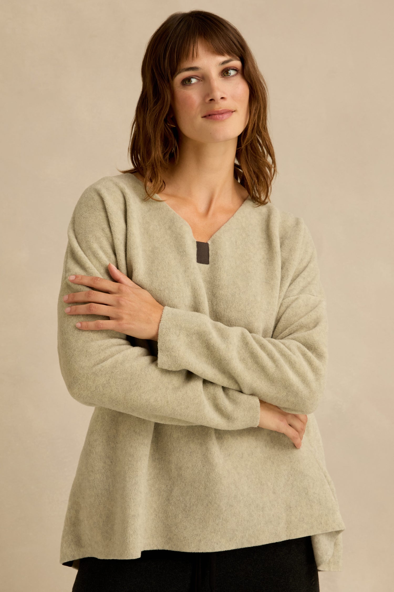 A person with shoulder-length brown hair wearing a light beige Cream Marl V Neck Fleece Tunic stands with arms crossed in front of a neutral background.