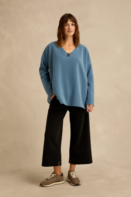 A woman stands on a plain background wearing the Dusty Blue V Neck Fleece Tunic, styled with wide black pants and brown sneakers, one hand casually in her pocket.