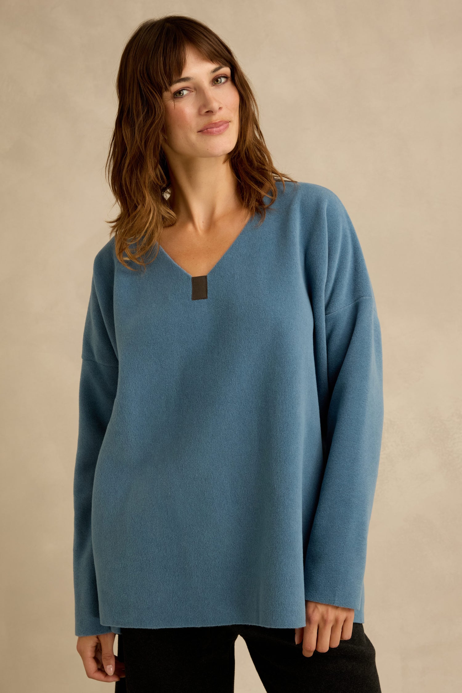 A woman models the Dusty Blue V Neck Fleece Tunic, a loose-fit sweater with a small brown tag at the neckline, set against a beige background.