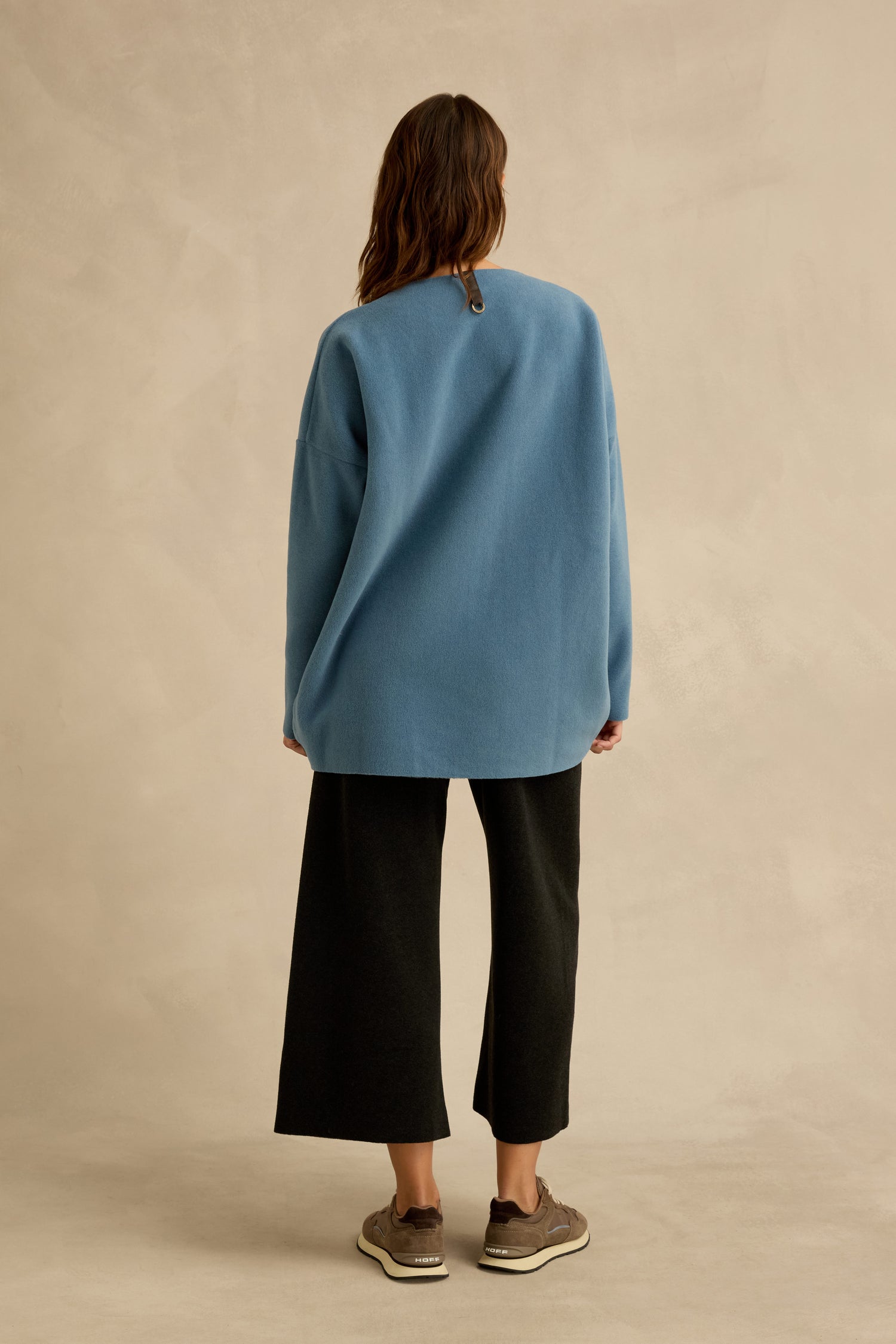 A person stands facing away, wearing the Dusty Blue V Neck Fleece Tunic with a loose fit, paired with black wide-leg pants and brown sneakers against a neutral backdrop.