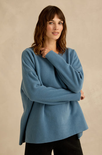 A woman with wavy brown hair stands against a neutral beige background, wearing a loose fit Dusty Blue V Neck Fleece Tunic and black pants.