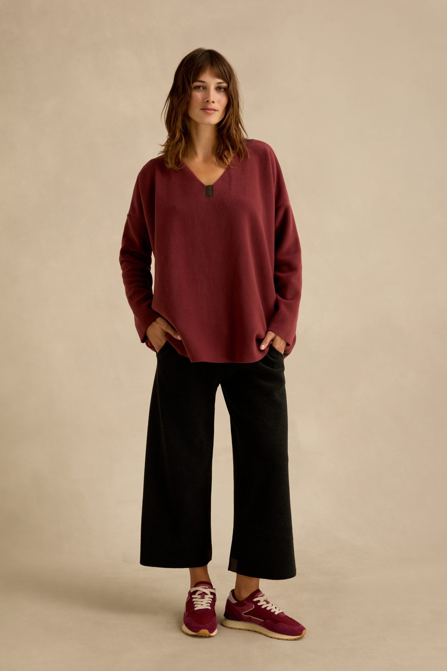 A woman stands against a plain background, wearing a loose Dusty Pink V Neck Fleece Tunic, black wide-leg pants, and maroon sneakers, with her hands in her pockets.