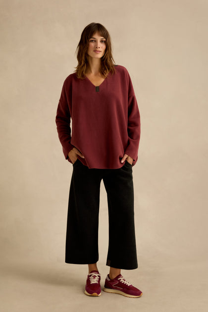 A woman stands against a plain background, wearing a loose Dusty Pink V Neck Fleece Tunic, black wide-leg pants, and maroon sneakers, with her hands in her pockets.
