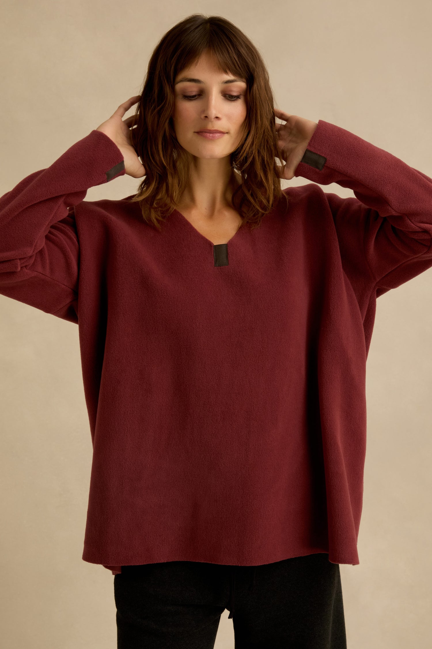 A woman stands against a neutral background wearing a loose-fitting Dusty Pink V Neck Fleece Tunic featuring dark accents at the collar and cuffs, paired with black pants.