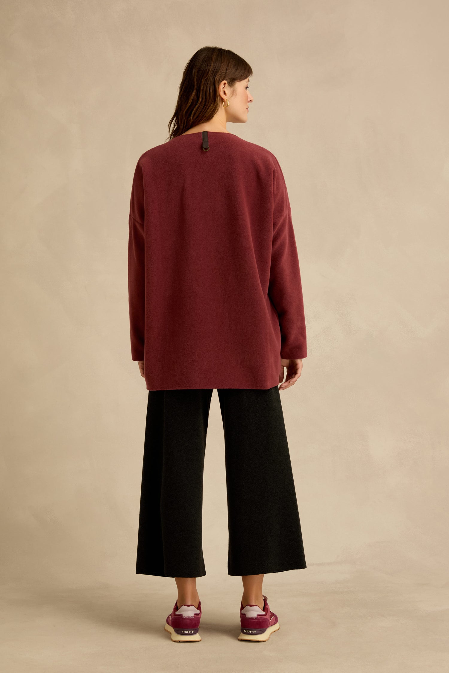 Person standing facing away, wearing a Henriette Steffensen Dusty Pink V Neck Fleece Tunic, black wide-leg pants, and maroon sneakers, against a beige background.
