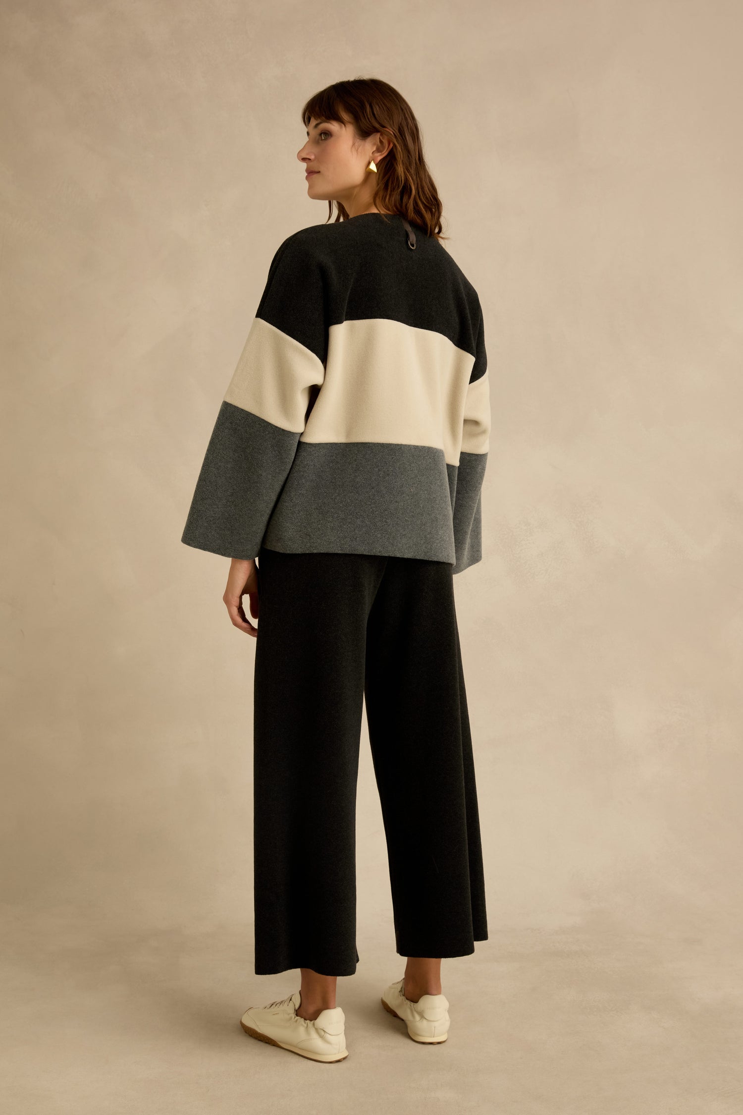 A person stands facing away, wearing a Colour Block Fleece Boxy Top by Henriette Steffensen in black, cream, and gray, paired with wide-leg black pants and white sneakers, against a neutral background.