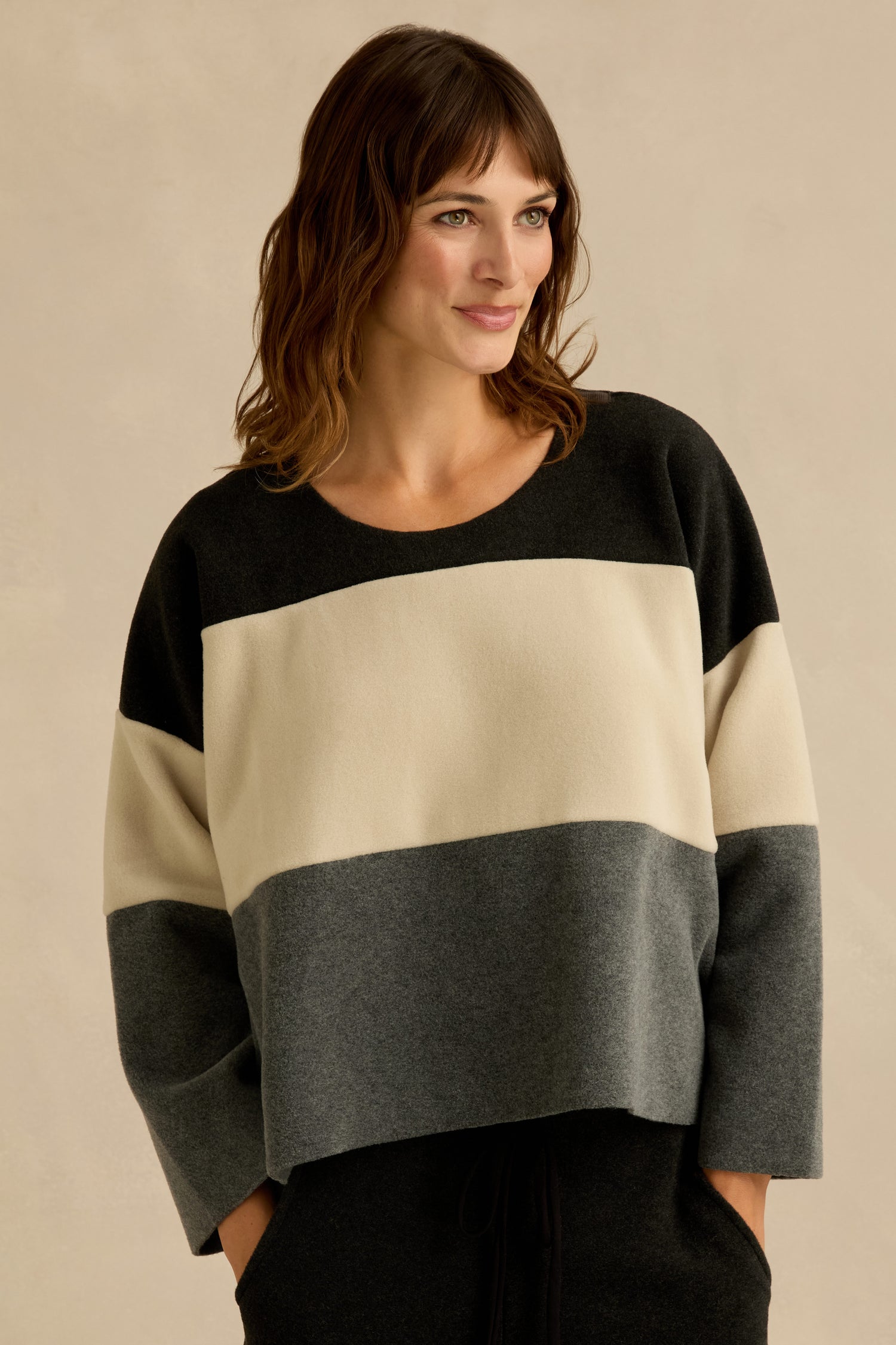 A woman wearing the Colour Block Fleece Boxy Top by Henriette Steffensen, featuring a boxy fit and wide horizontal color blocks in black, cream, and gray, stands against a plain beige background.