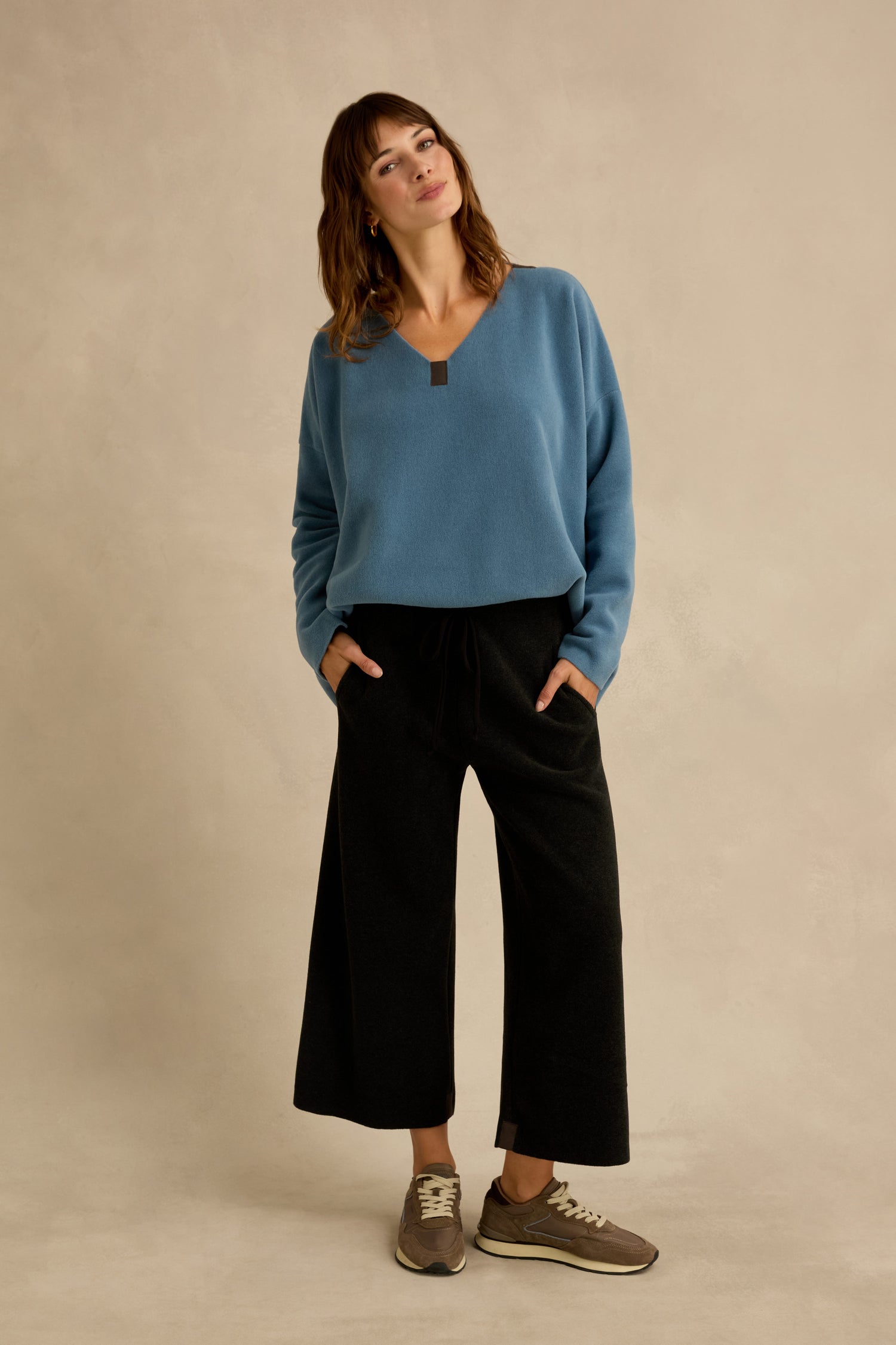 Standing against a beige background, the woman wears a blue sweater paired with cropped Straight Leg Fleece Trousers in black and brown sneakers, her hands in her pockets and head slightly tilted.