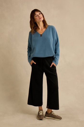 Standing against a beige background, the woman wears a blue sweater paired with cropped Straight Leg Fleece Trousers in black and brown sneakers, her hands in her pockets and head slightly tilted.