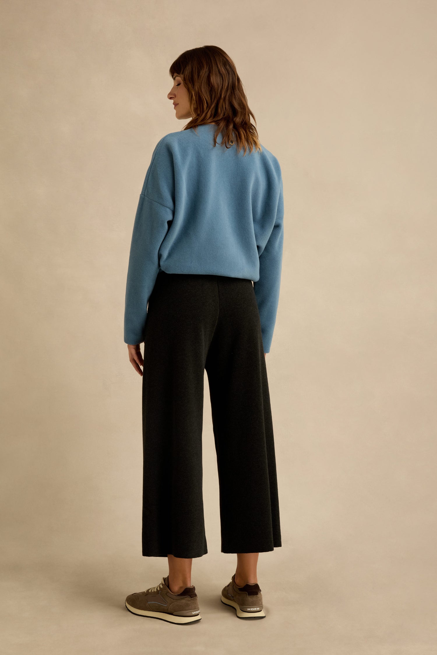 A person stands facing away, wearing a light blue sweater, Straight Leg Fleece Trousers in a cropped black wide-leg style, and brown sneakers against a plain beige background.