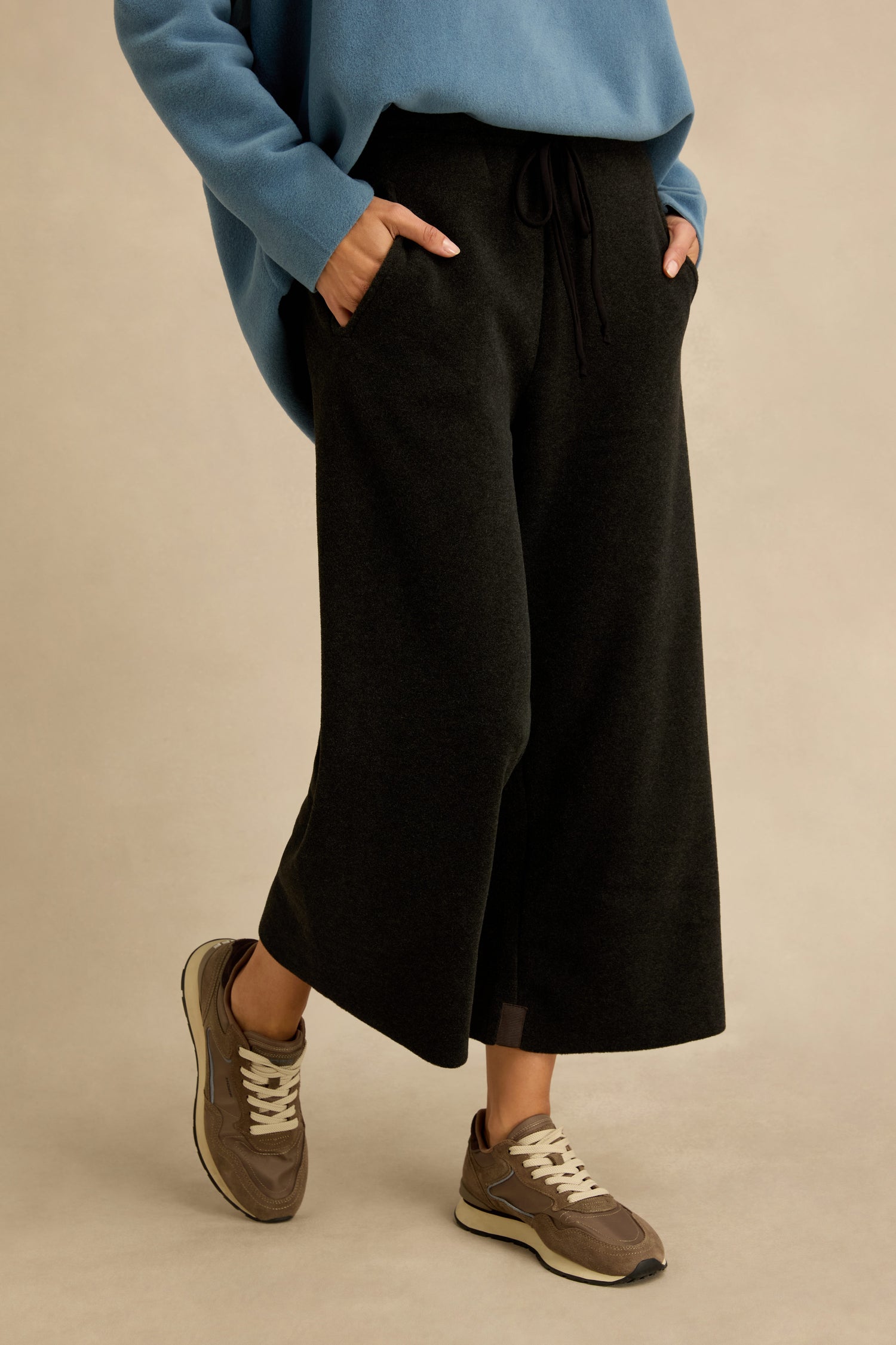 Wearing the Straight Leg Fleece Trousers in black, along with a blue sweater and brown sneakers, a person stands with hands in pockets against a plain beige background.