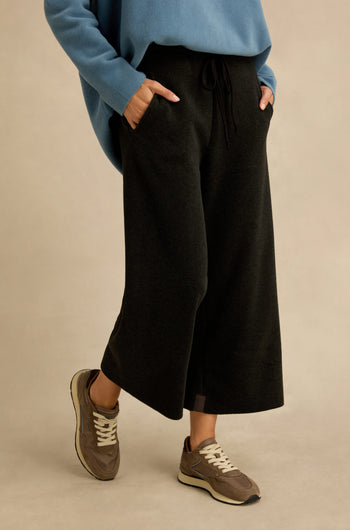 Wearing the Straight Leg Fleece Trousers in black, along with a blue sweater and brown sneakers, a person stands with hands in pockets against a plain beige background.