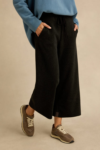 Wearing the Straight Leg Fleece Trousers in black, along with a blue sweater and brown sneakers, a person stands with hands in pockets against a plain beige background.