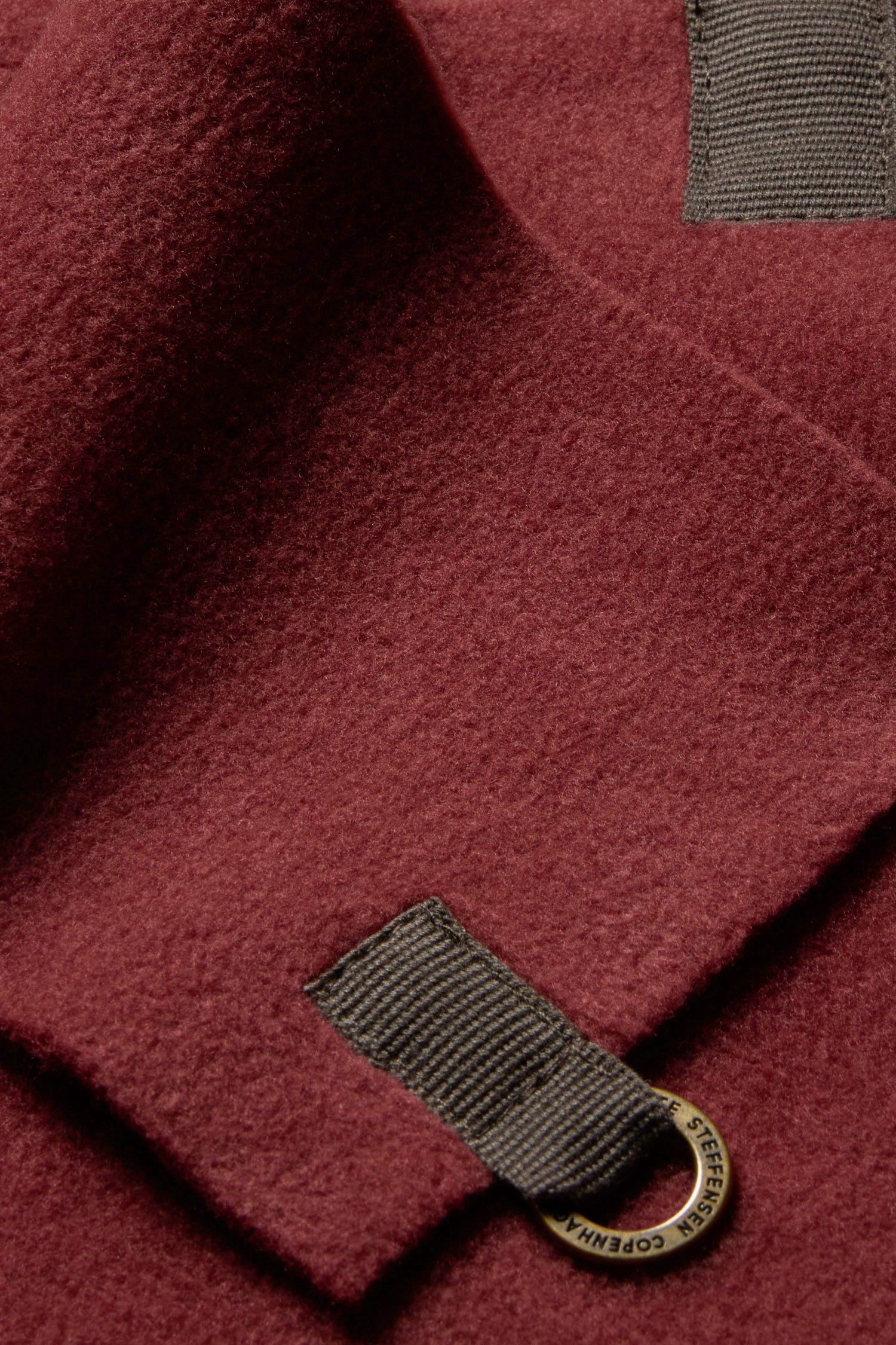 Close-up of a folded burgundy Triangle Scarf with dark gray tabs and a circular metal tag attached to one tab, reminiscent of the signature detailing found on Henriette Steffensen’s triangle scarves and fleece scarves.