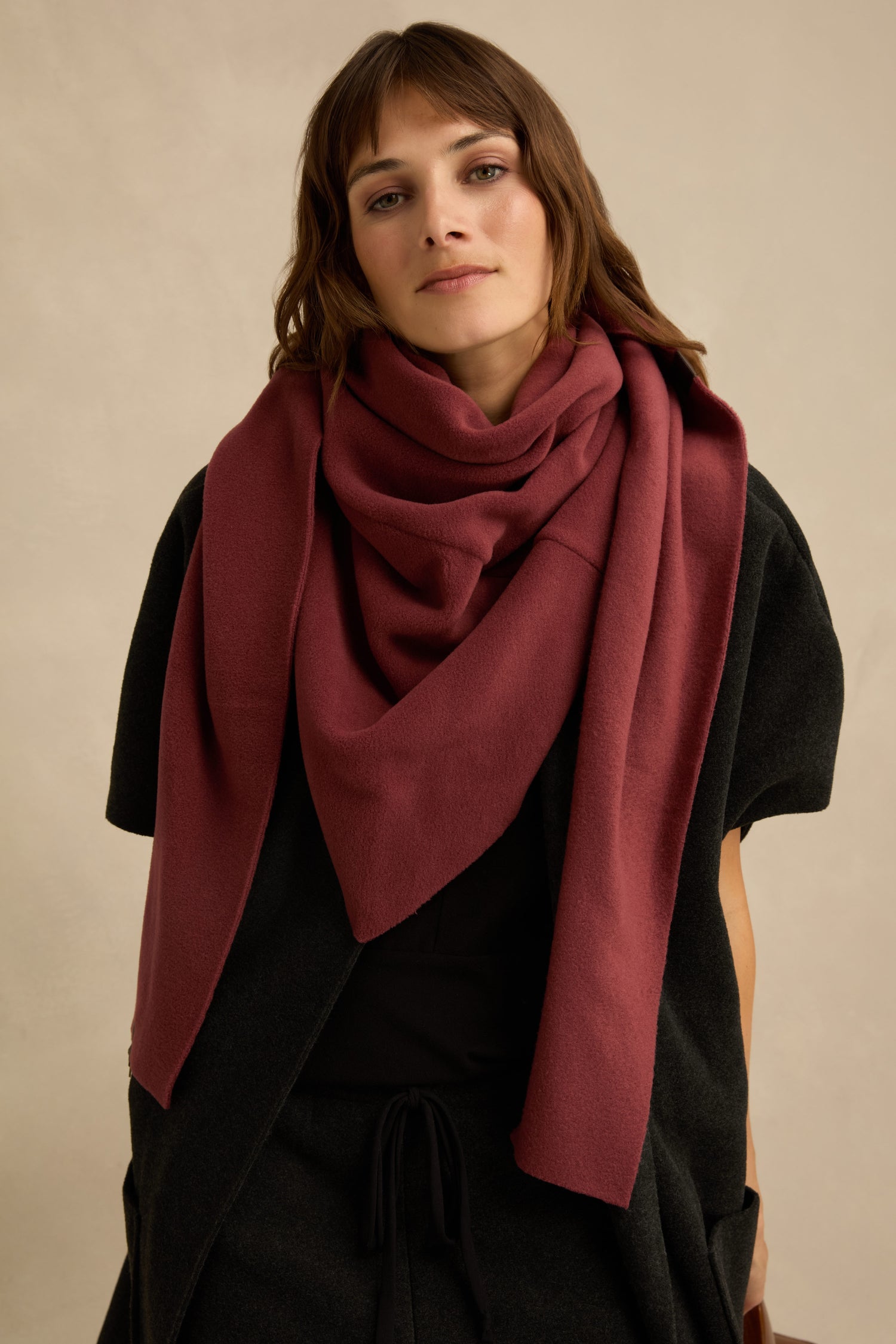A woman wears the Triangle Scarf in large burgundy fleece over a black outfit, standing against a neutral background and looking at the camera with a neutral expression.