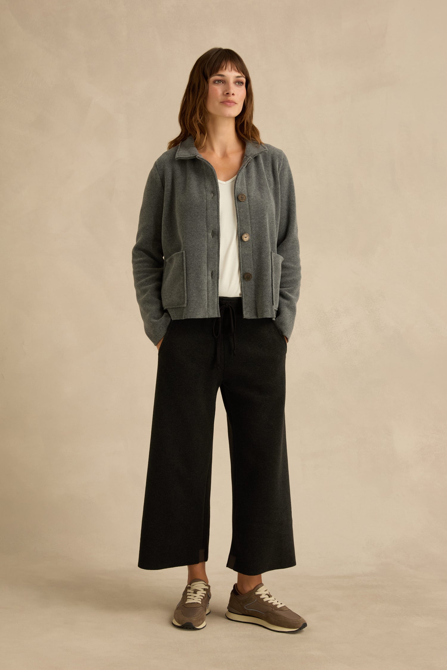 A woman stands against a beige background wearing a Grey Fleece Fitted Jacket, white top, black wide-leg pants, and gray sneakers, with her hands in her pockets.
