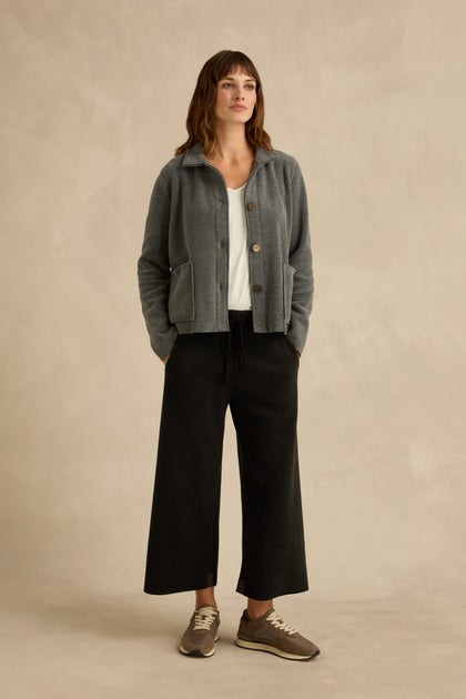 A woman stands against a beige background wearing a Grey Fleece Fitted Jacket, white top, black wide-leg pants, and gray sneakers, with her hands in her pockets.