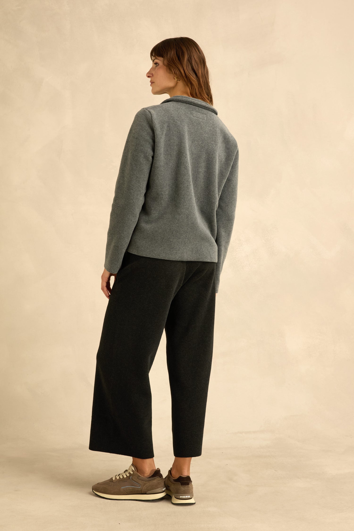 A person stands facing away, wearing a Grey Fleece Fitted Jacket, black wide-leg pants, and taupe sneakers against a beige background.