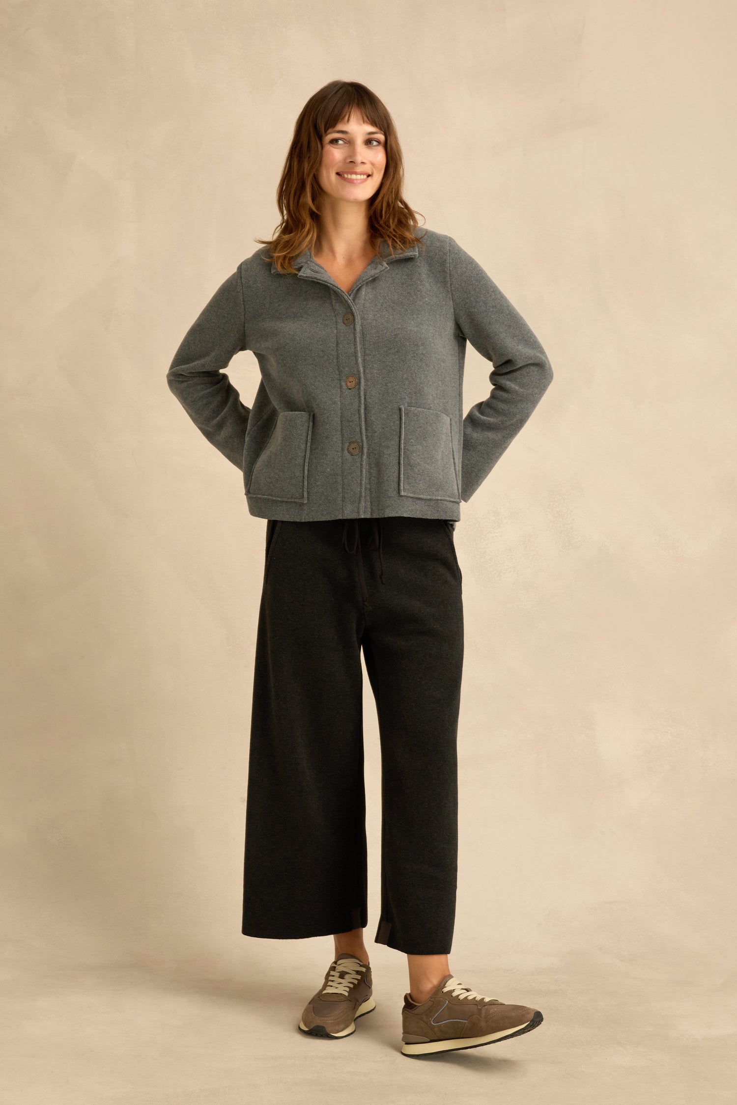A person stands smiling against a beige background, wearing the Grey Fleece Fitted Jacket from Henriette Steffensen, wide-leg black pants, and brown sneakers.