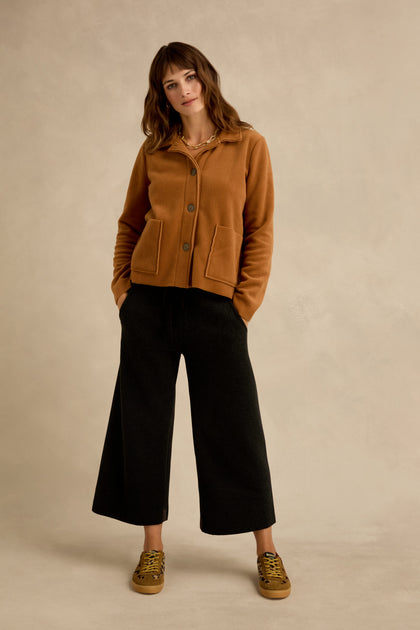 Woman standing against a neutral background wearing the Rust Fleece Fitted Jacket, wide-leg black pants, and brown sneakers, with her hands in her pockets.