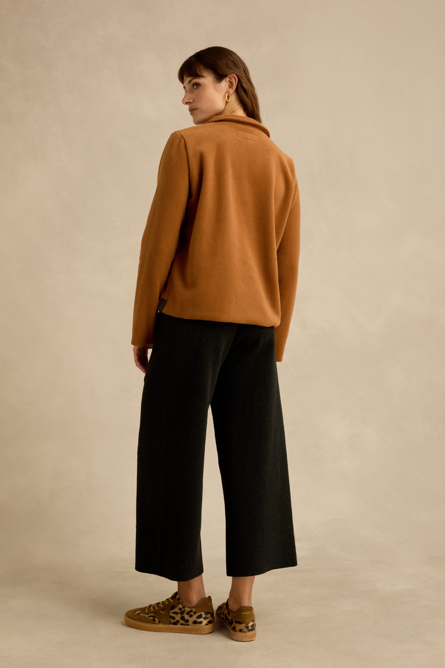 A person stands facing away, wearing the Rust Fleece Fitted Jacket, black wide-leg pants, and leopard print shoes against a neutral background.