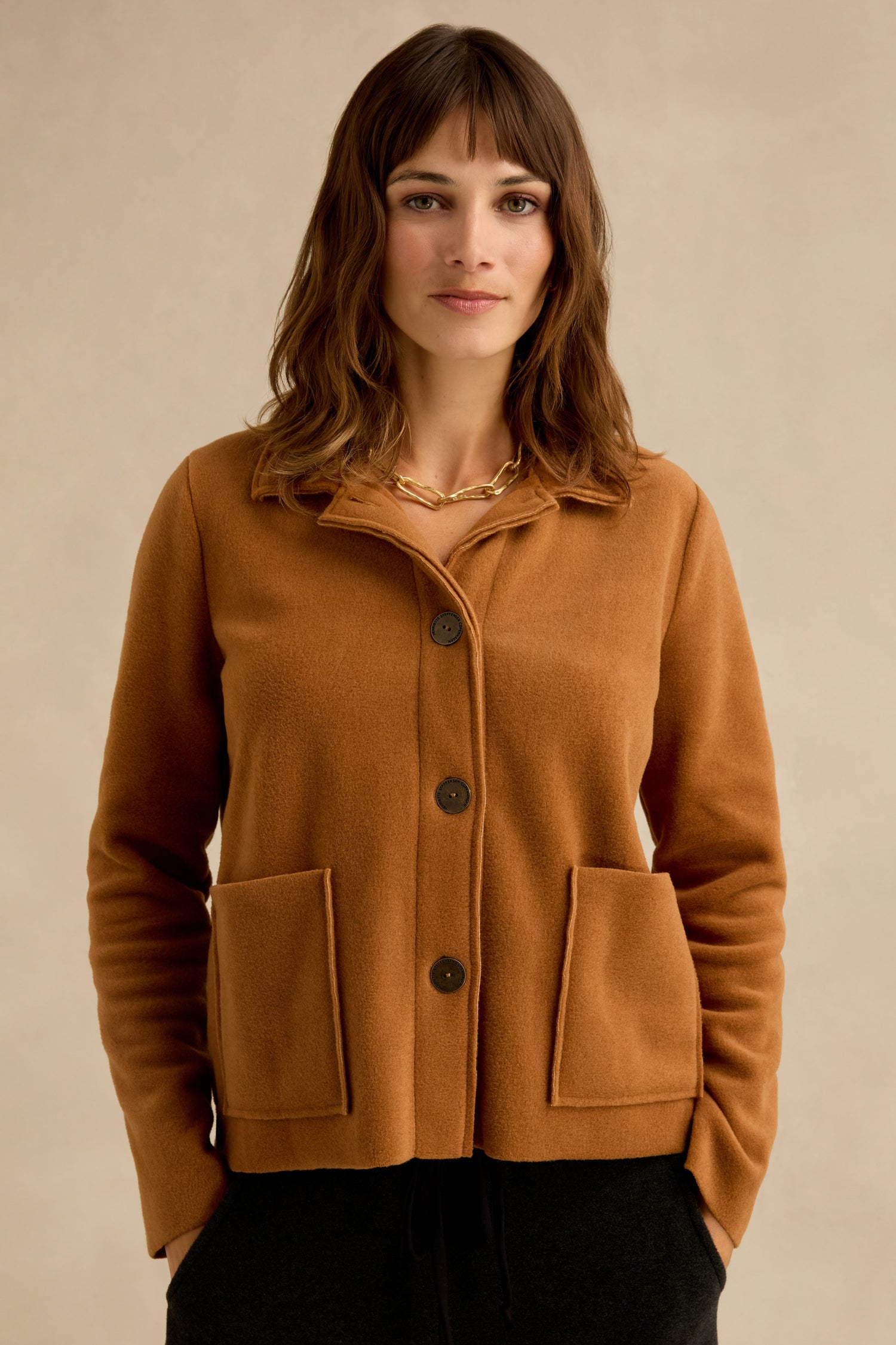 Woman standing against a neutral background, wearing a Rust Fleece Fitted Jacket with large front pockets and a gold chain necklace.