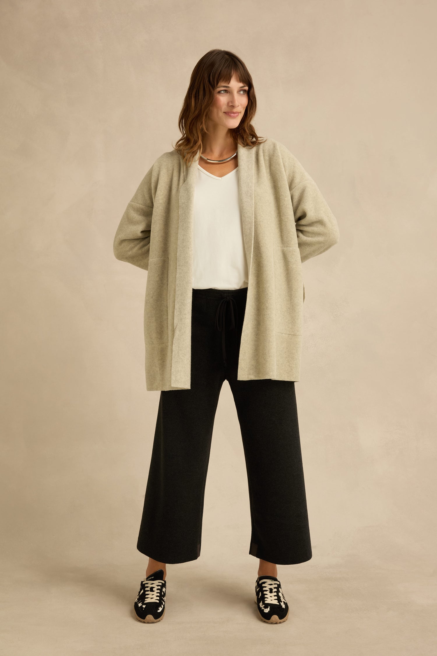 A woman stands against a neutral background wearing the Cream Marl Fleece Cardigan over a white top, paired with black wide-leg pants and black sneakers with white soles.