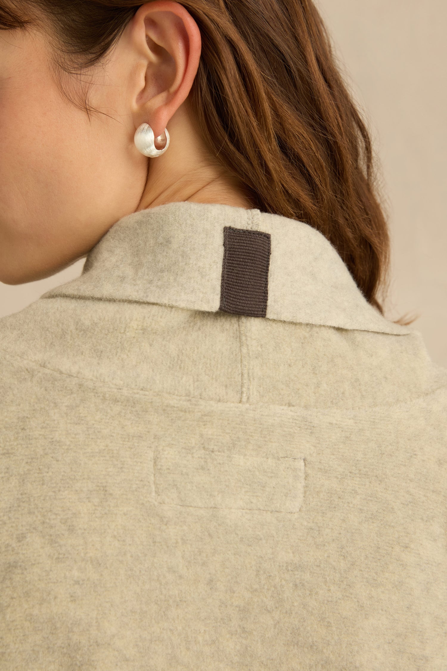 Close-up of a person’s ear wearing a silver hoop earring, highlighting the back collar detail of the Cream Marl Fleece Cardigan, which features a dark fabric loop and a relaxed fit.