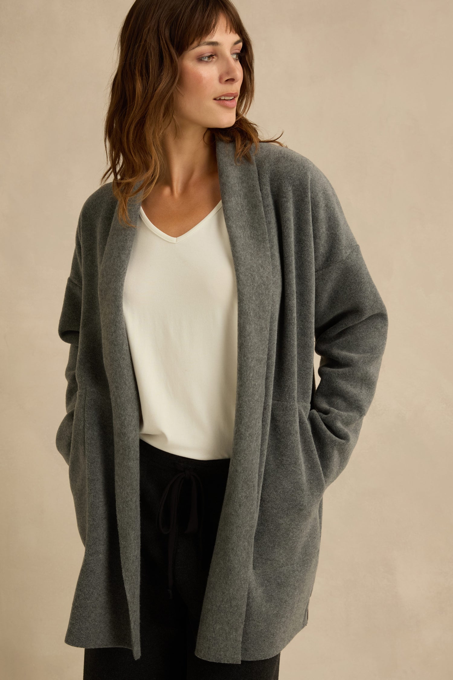 A woman wearing the Grey Fleece Cardigan layered over a white top and black pants stands with her hands in the pockets against a neutral background.