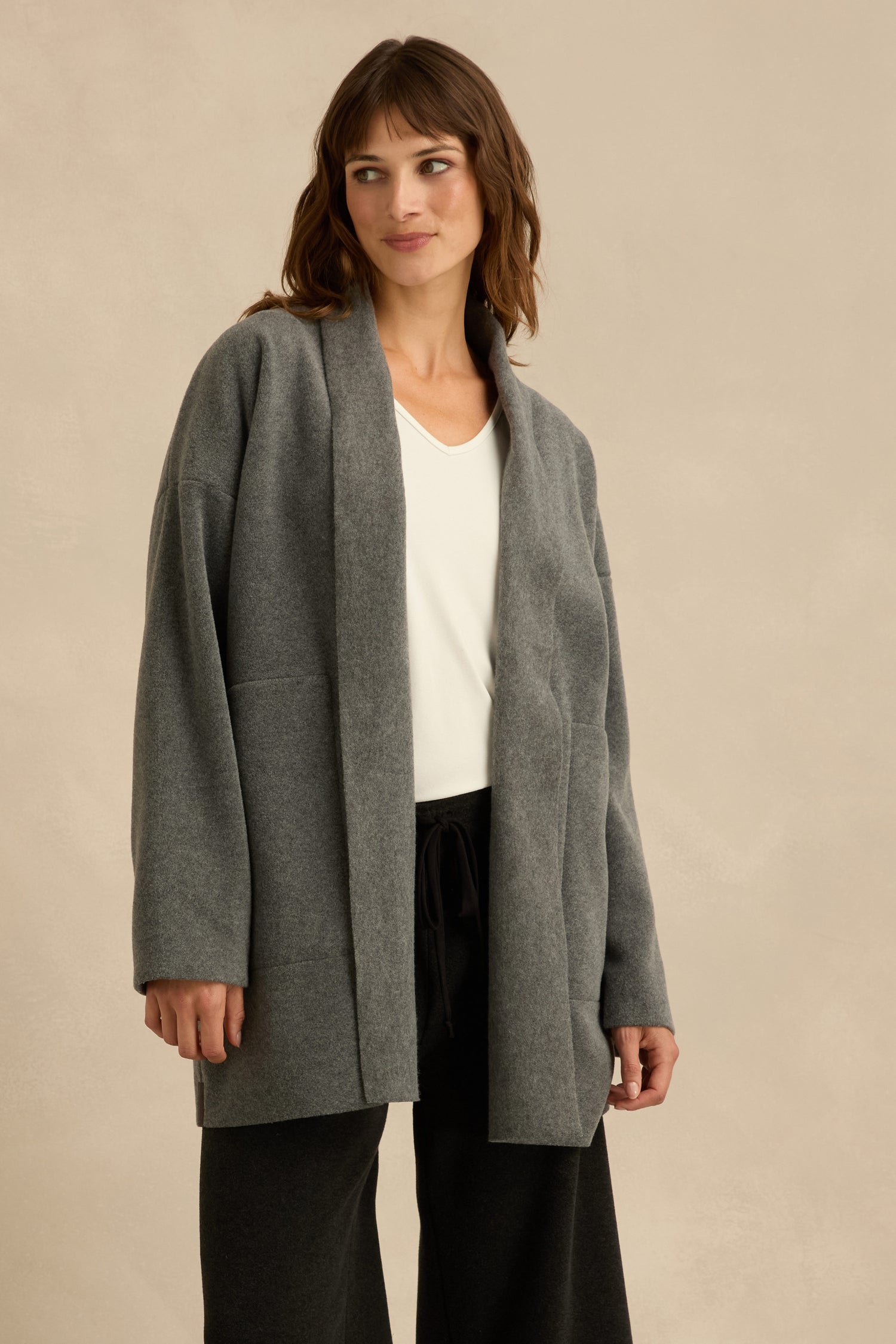 A woman stands against a beige background wearing the Grey Fleece Cardigan, a relaxed-fit, gray open-front style, layered over a white top and black pants.