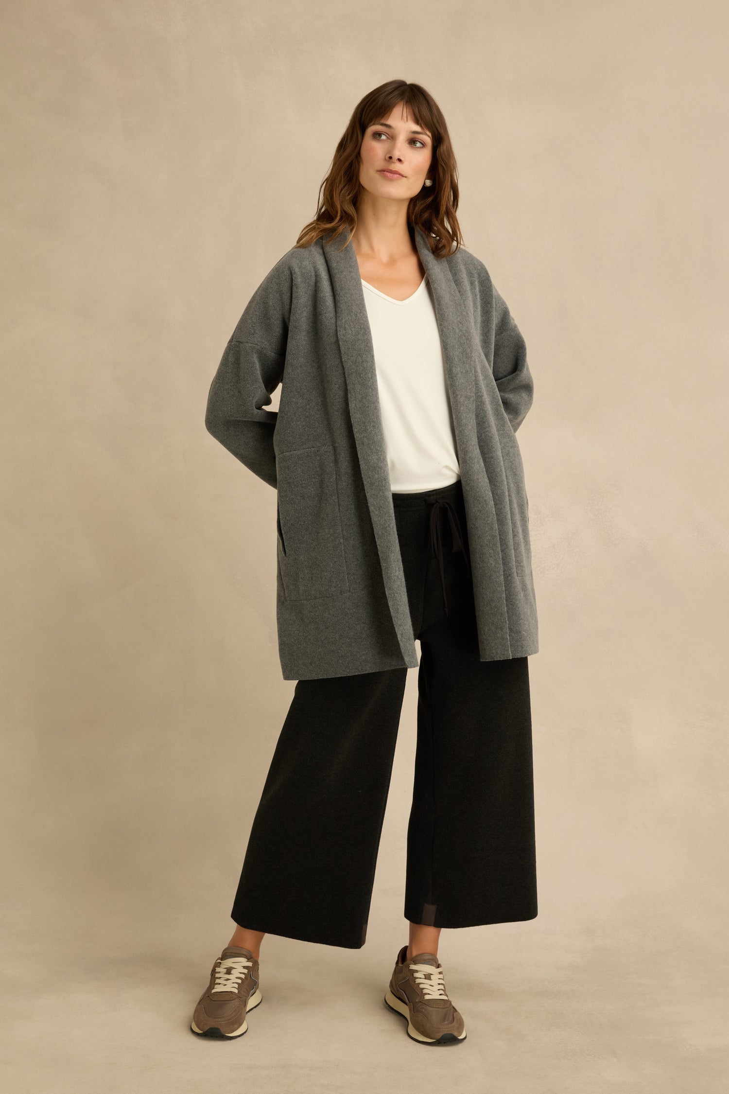 A woman stands against a neutral background, wearing a relaxed fit Grey Fleece Cardigan over a white V-neck top, paired with wide-leg black pants and beige sneakers, with her hands behind her back.