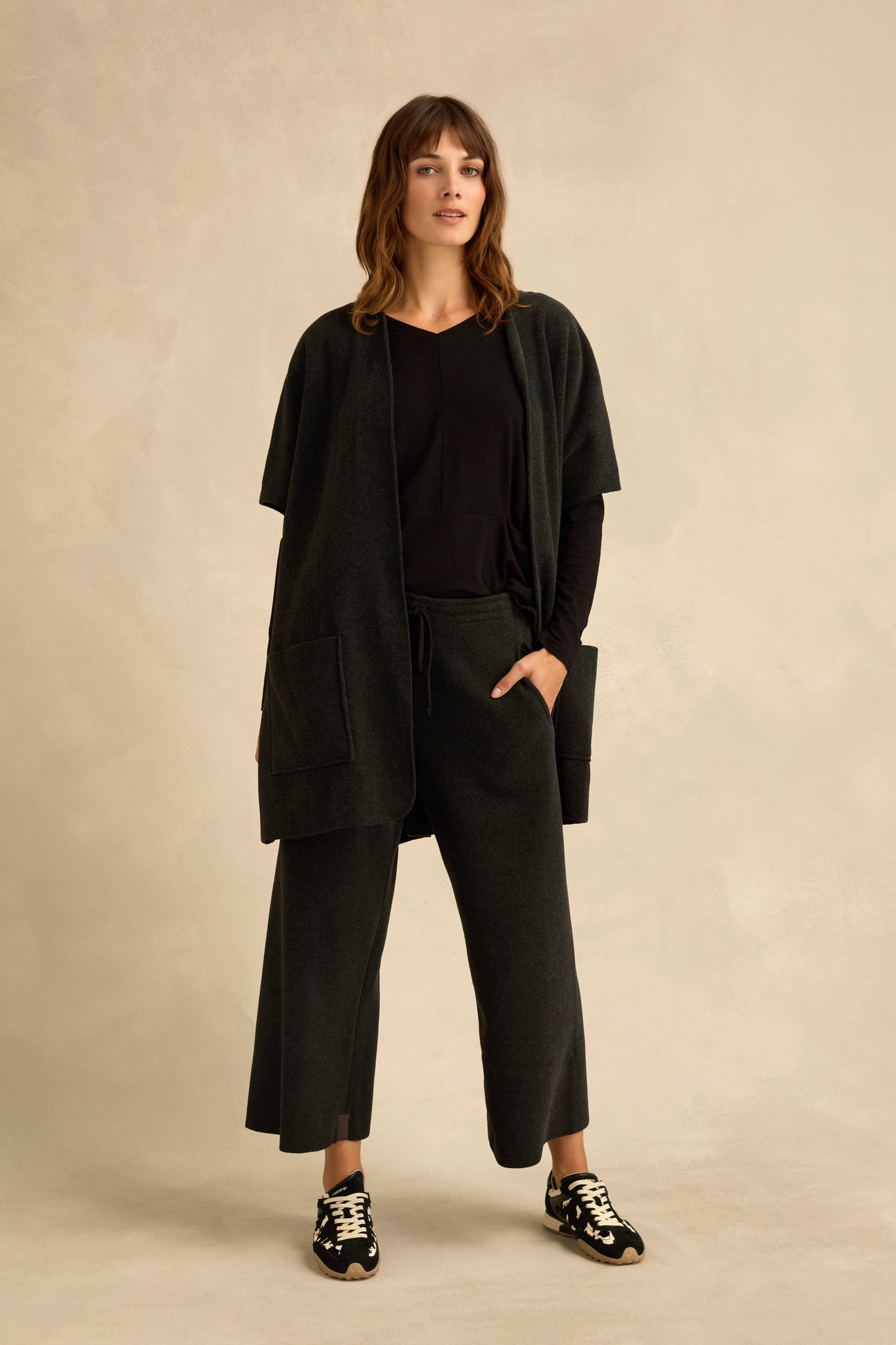 A woman stands against a beige background, wearing a short sleeve fleece gilet layered over a black top and matching wide-leg pants, completed with black sneakers featuring white soles.