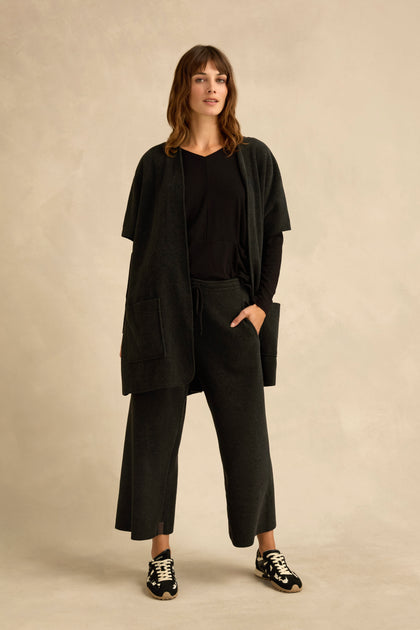 A woman stands against a beige background, wearing a short sleeve fleece gilet layered over a black top and matching wide-leg pants, completed with black sneakers featuring white soles.