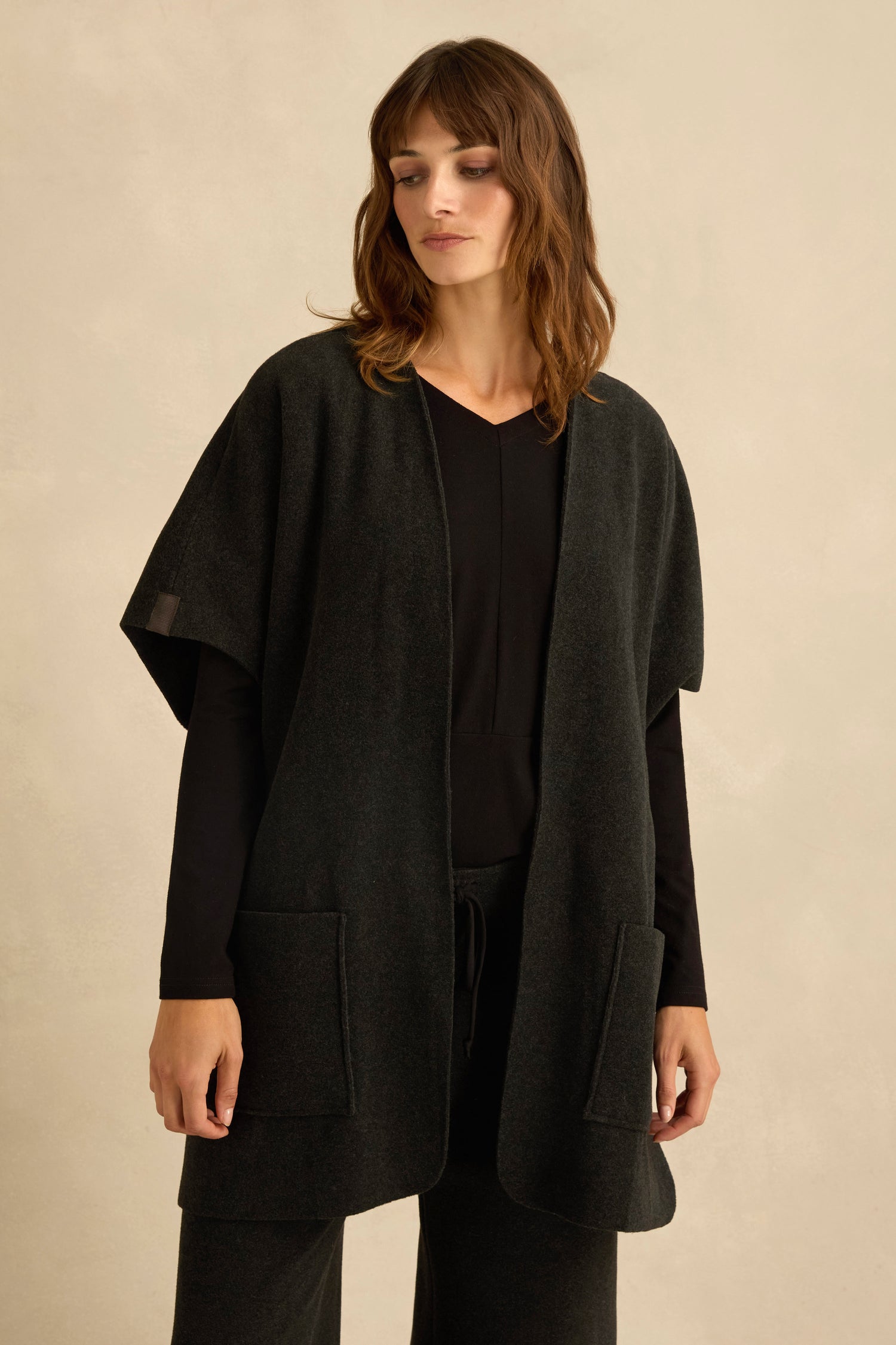 A woman stands against a plain beige background wearing a dark Short Sleeve Fleece Gilet with kimono-style sleeves and large front pockets, layered over a black long-sleeved top and matching pants.